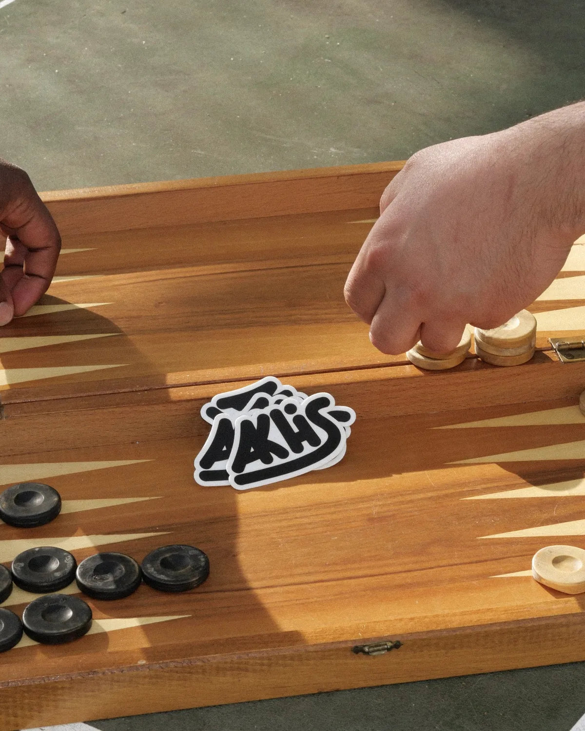 Zwei Hände spielen Tavla, auf dem Brett klebt ein Akhis-Sticker.